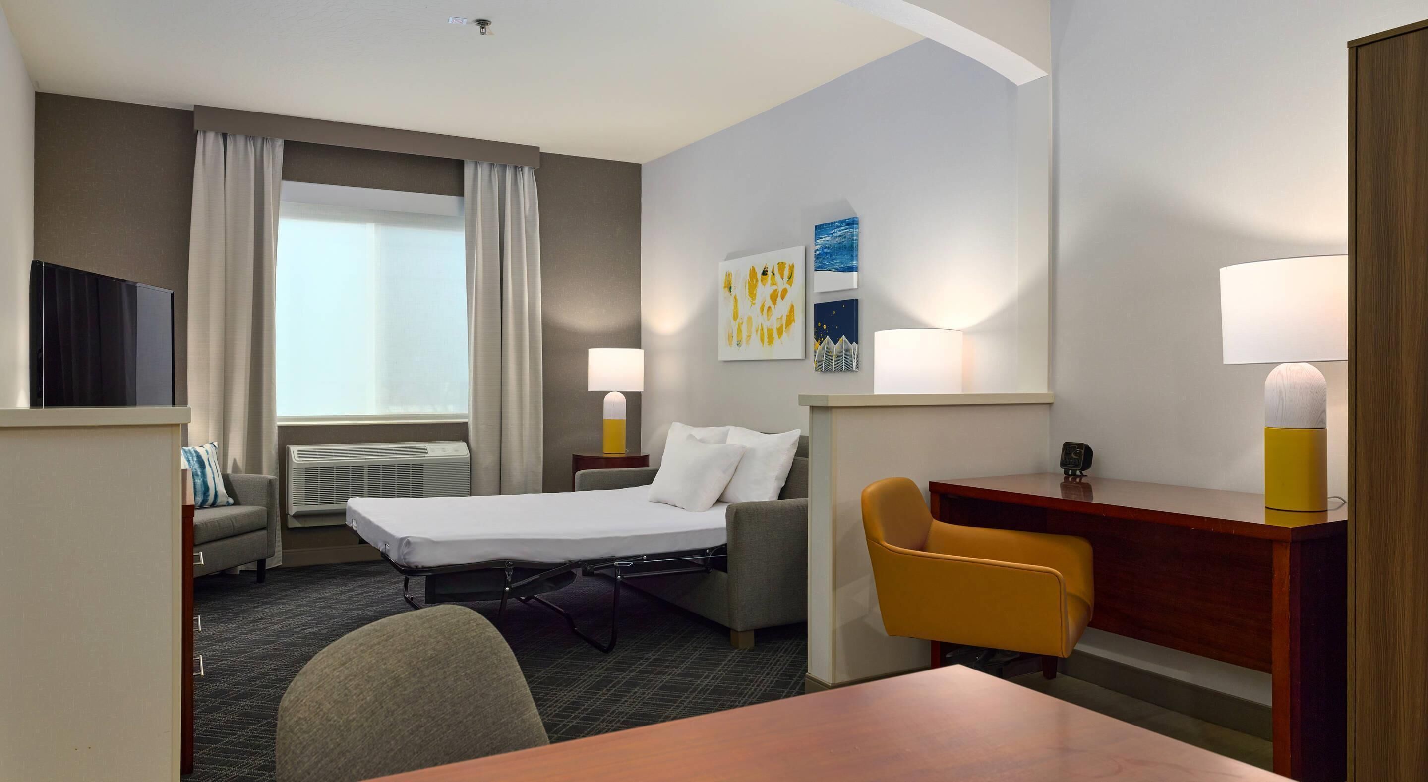 Una habitación de hotel con cama, escritorio, silla, televisión y ventana con cortinas.