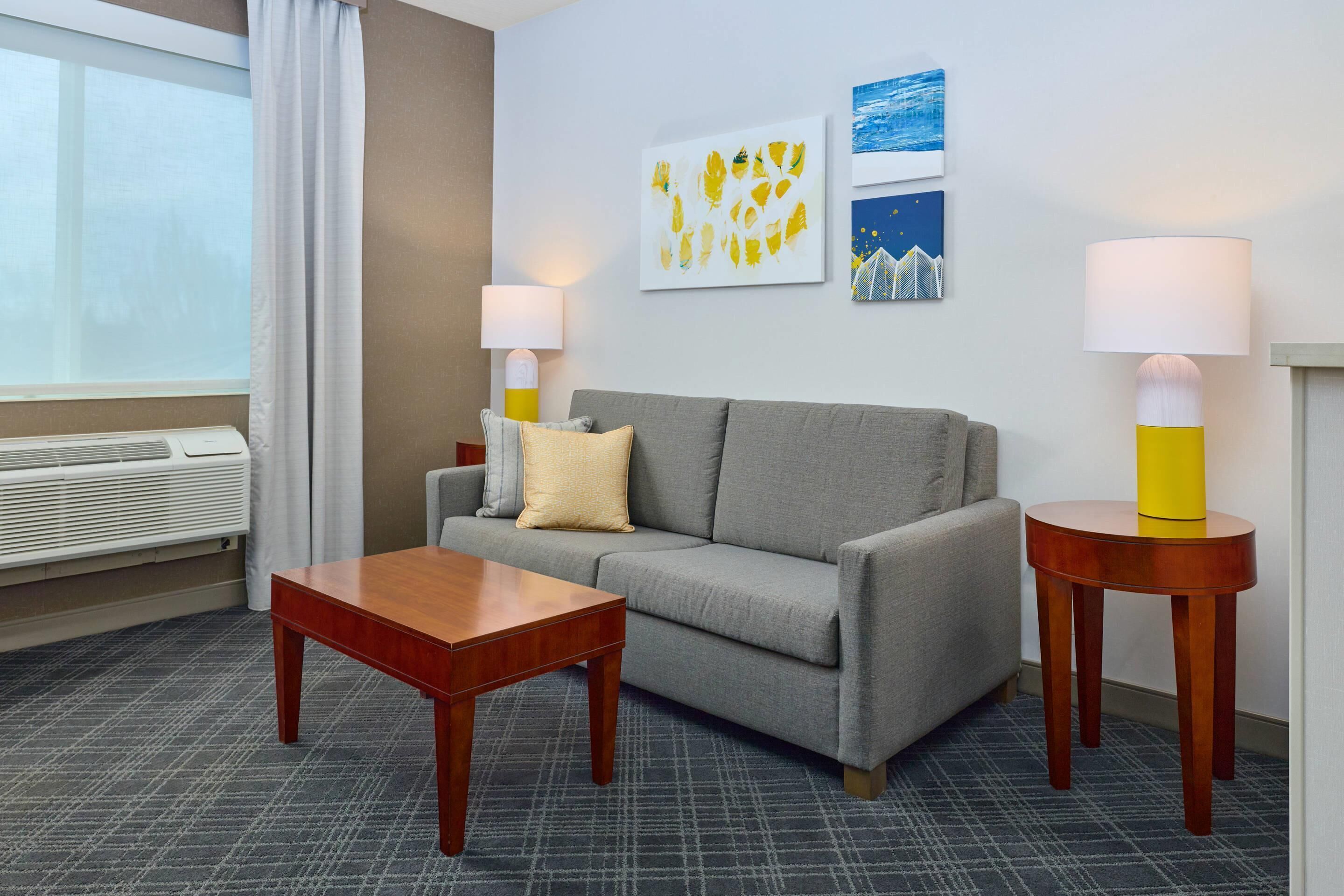 A hotel room with a grey sofa, a wooden coffee table, two bedside lamps, and artwork on the wall.