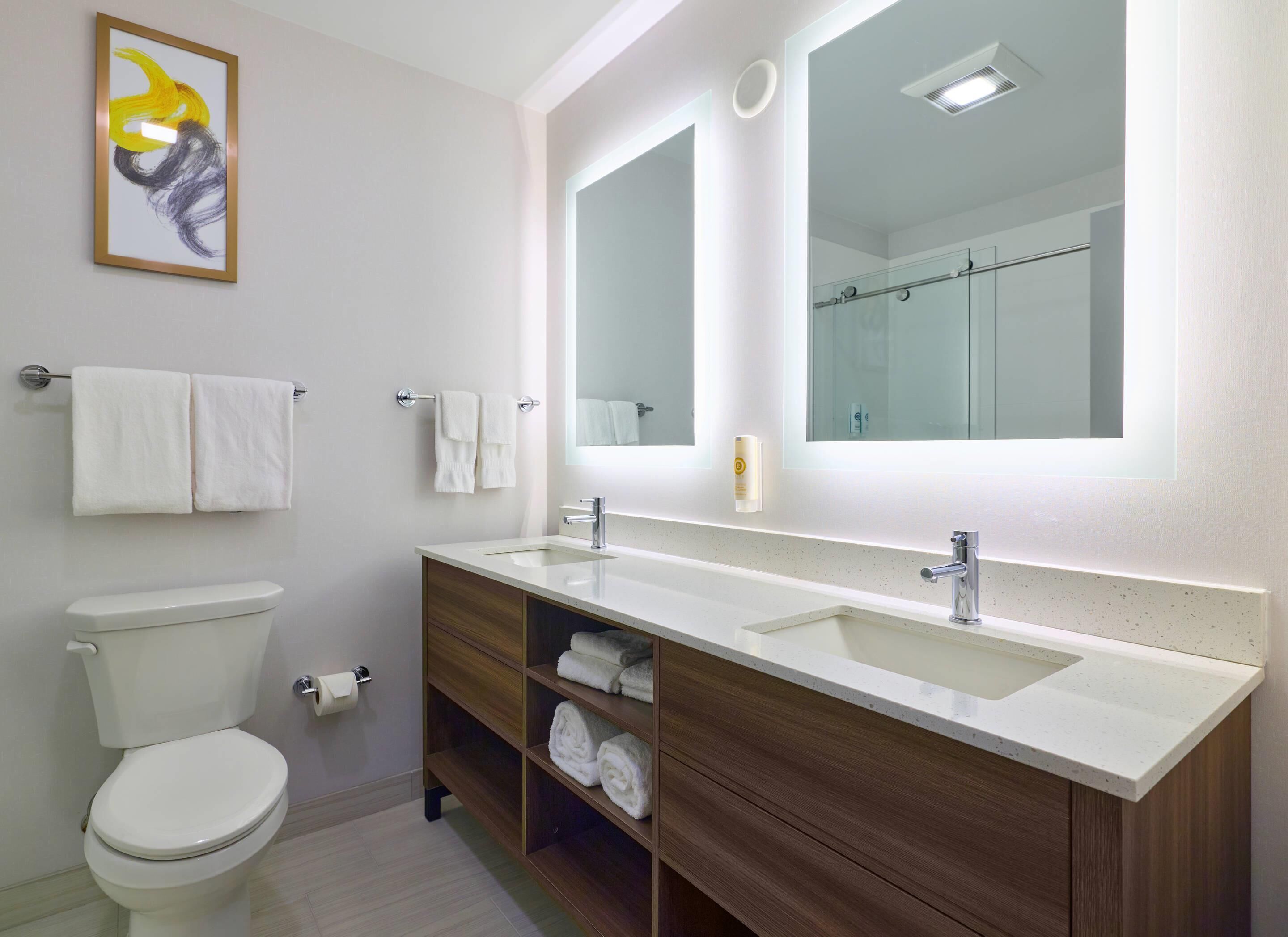 A modern bathroom with a toilet, two sinks, a large mirror, and a framed artwork above the sink.