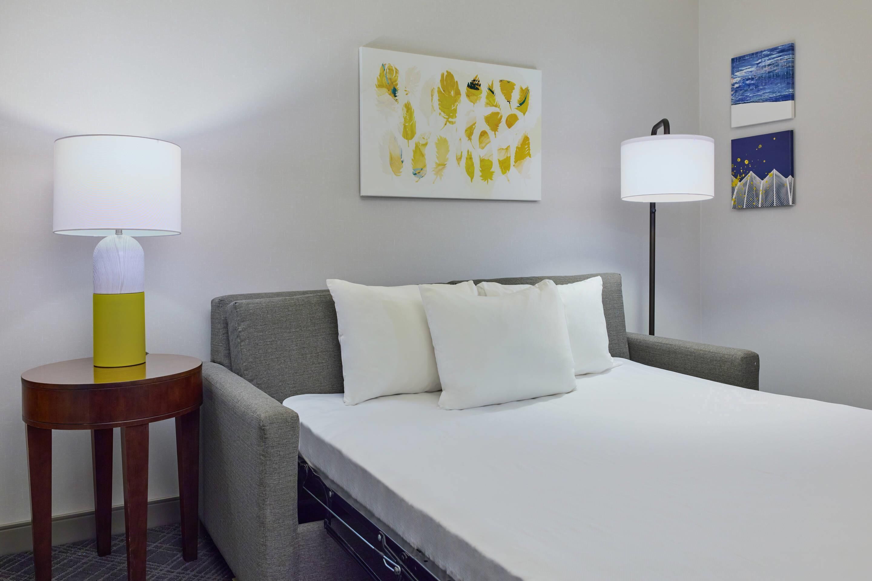 Habitación de hotel con cama, dos lámparas de noche, una mesita con lámpara y dos obras de arte en la pared.