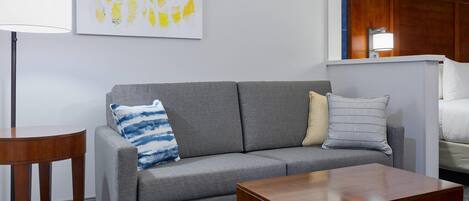 A modern hotel room with a grey sofa, two wooden side tables, a patterned rug, and a painting of yellow leaves on the wall.