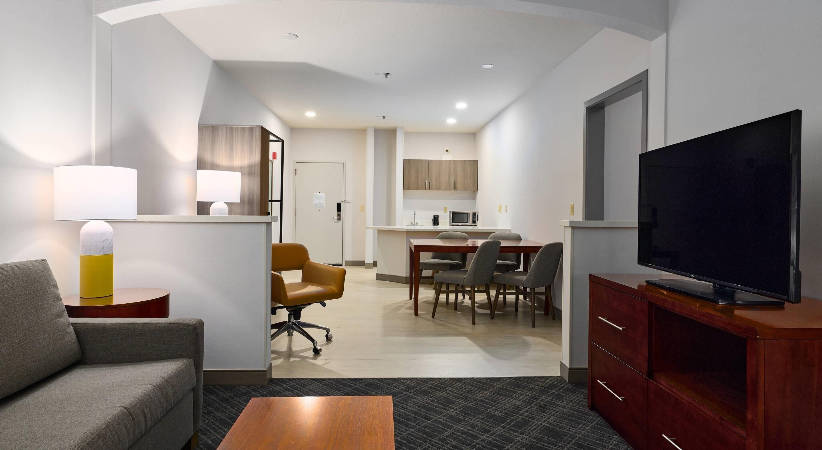 A hotel room with a sofa, a TV on a wooden cabinet, a dining area with chairs and a table, and a kitchen with cabinets and a microwave.