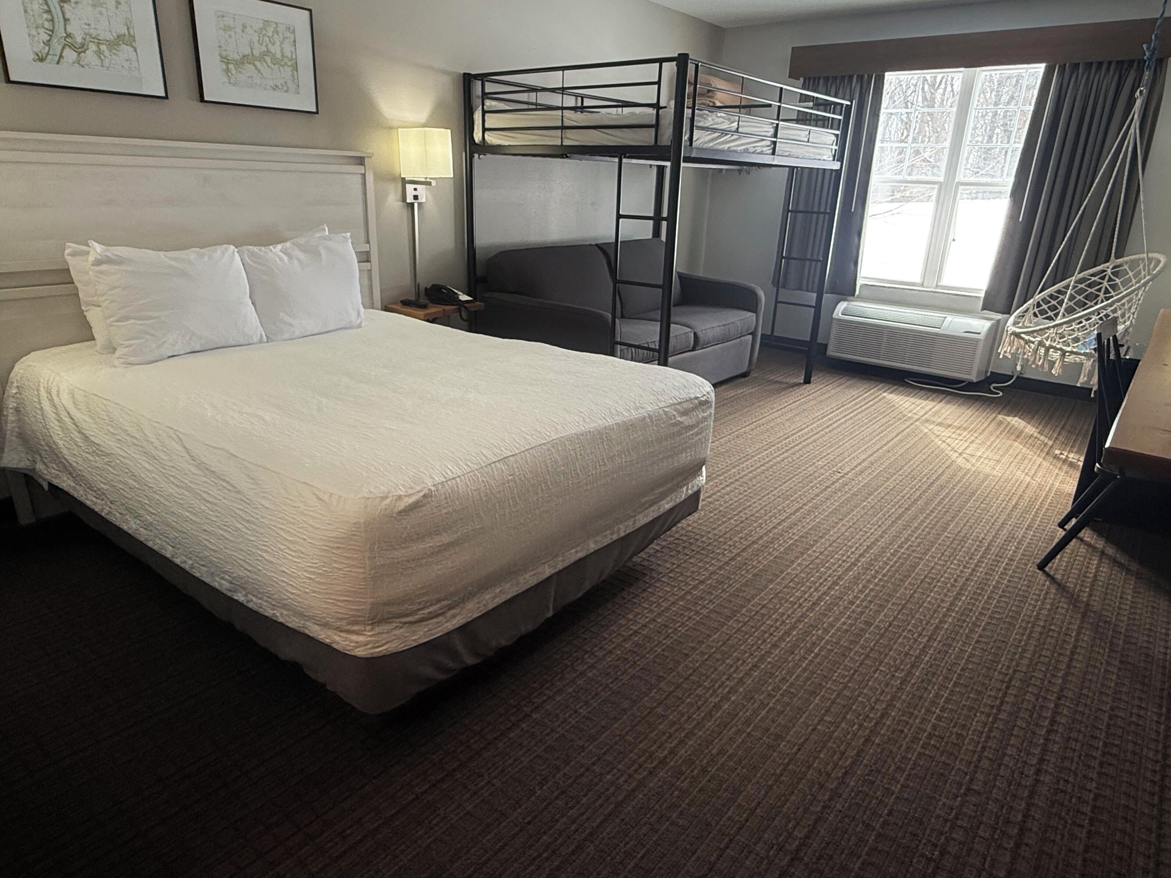 A hotel room with a bunk bed, a single bed, a desk, a chair, a lamp, and a window with curtains.