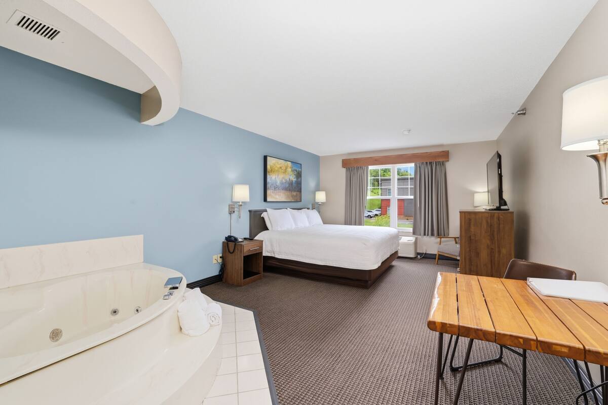 Ein Hotelzimmer mit Bett, Schreibtisch, Stuhl, Schreibtischlampe, Nachttisch, Lampe, einem Gemälde, einem Fenster mit Vorhängen und Blick ins Freie.