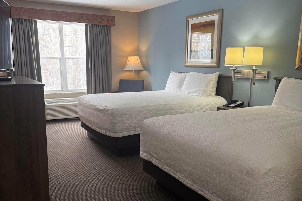 A hotel room with two beds, a desk, a lamp, and a window with curtains.