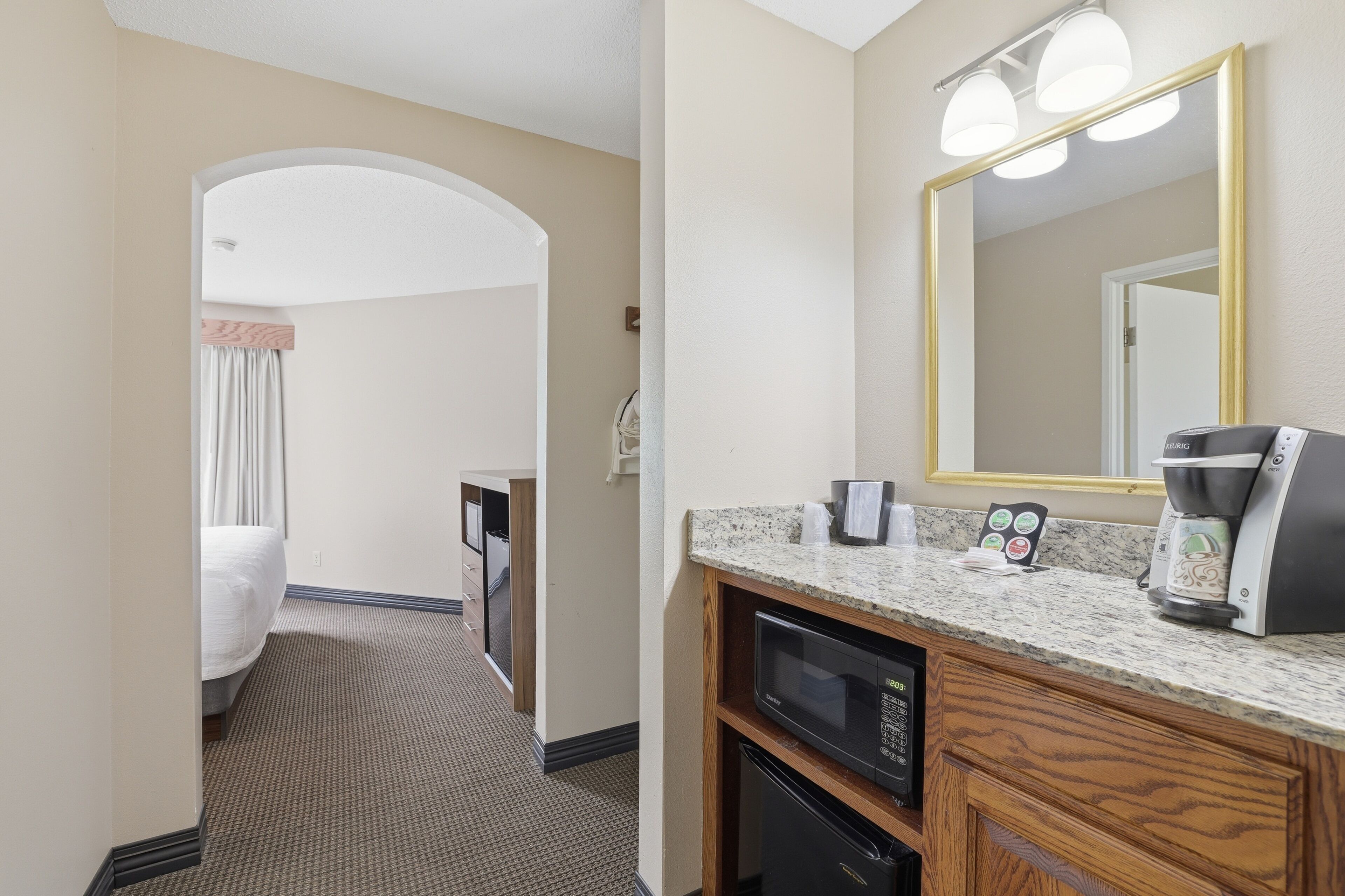 A hotel room with a kitchenette, a large mirror, a coffee maker, and a bed with a white comforter.