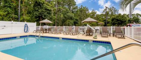 A swimming pool with lounge chairs and umbrellas, surrounded by a fence and trees.