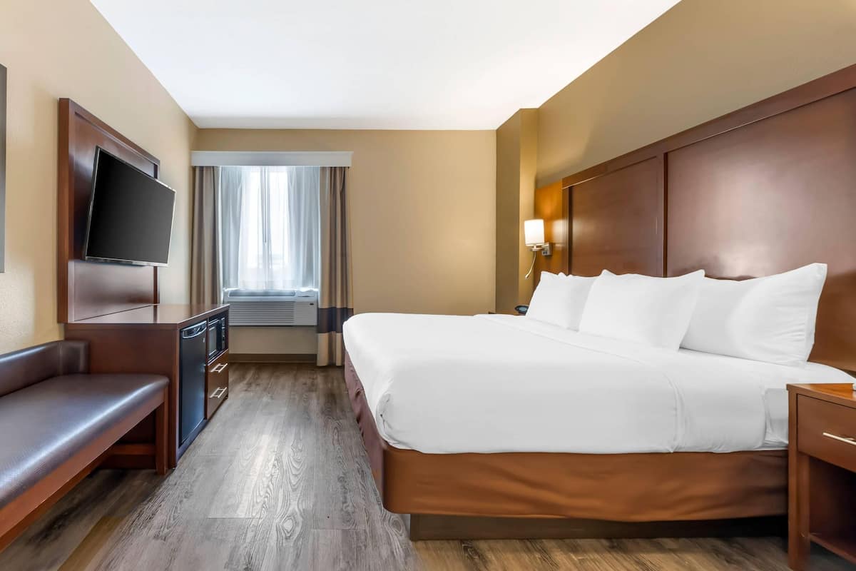 A hotel room with a bed, a TV, a desk, a bench, and a window with curtains.