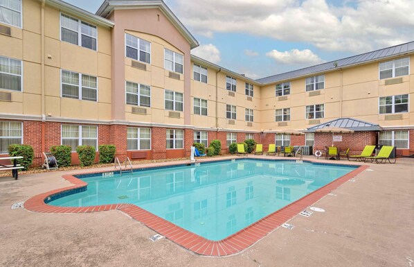 Uma área de piscina com uma piscina retangular, rodeada por um deck de tijolos, e adjacente a um edifício alto com janelas amplas e telhado plano.