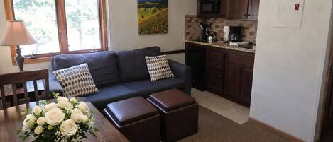 Un espace de vie compact comprenant une table à manger, un canapé, des poufs, un ventilateur de plafond, un coin cuisine et une fenêtre avec des rideaux.