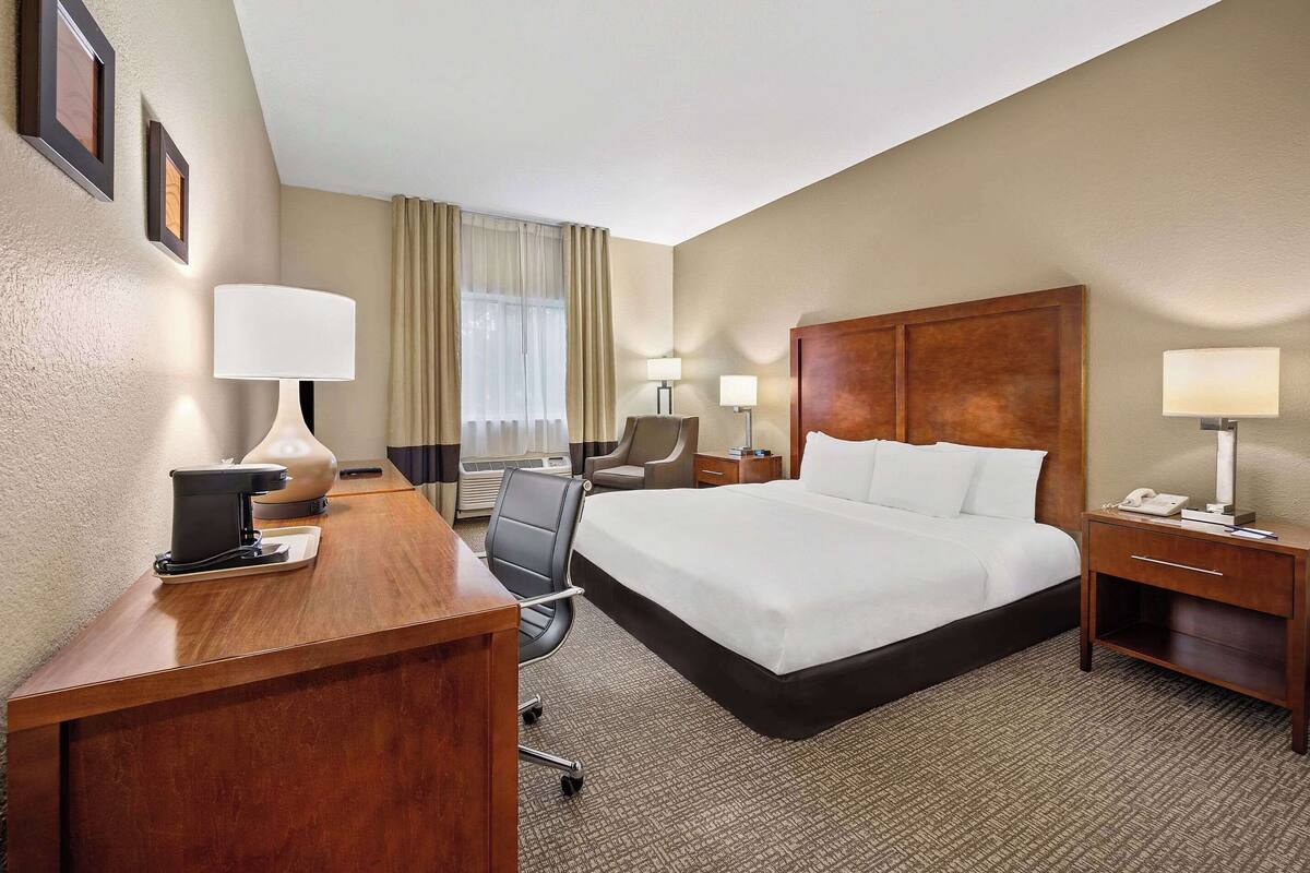 A hotel room with a large bed, a desk with a lamp, a chair, and a nightstand with a phone.