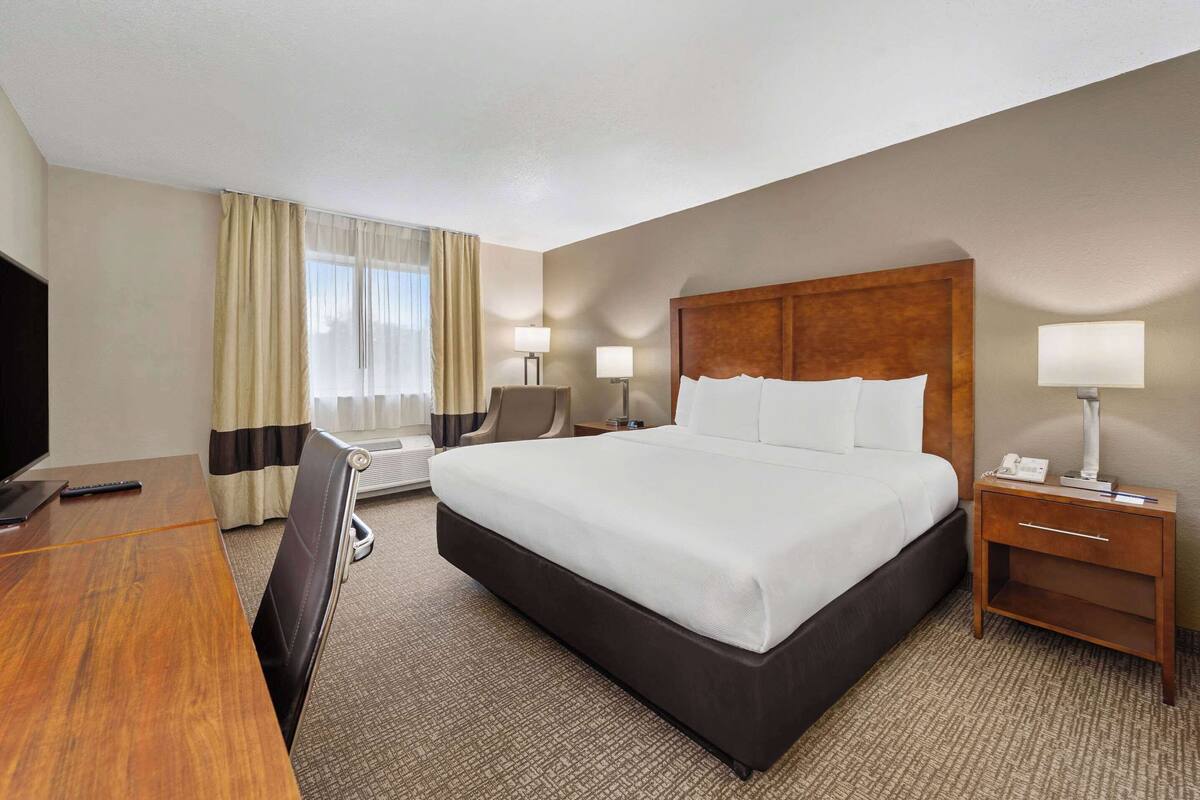 A hotel room with a large bed, a desk, two chairs, a TV, and a nightstand with a lamp.