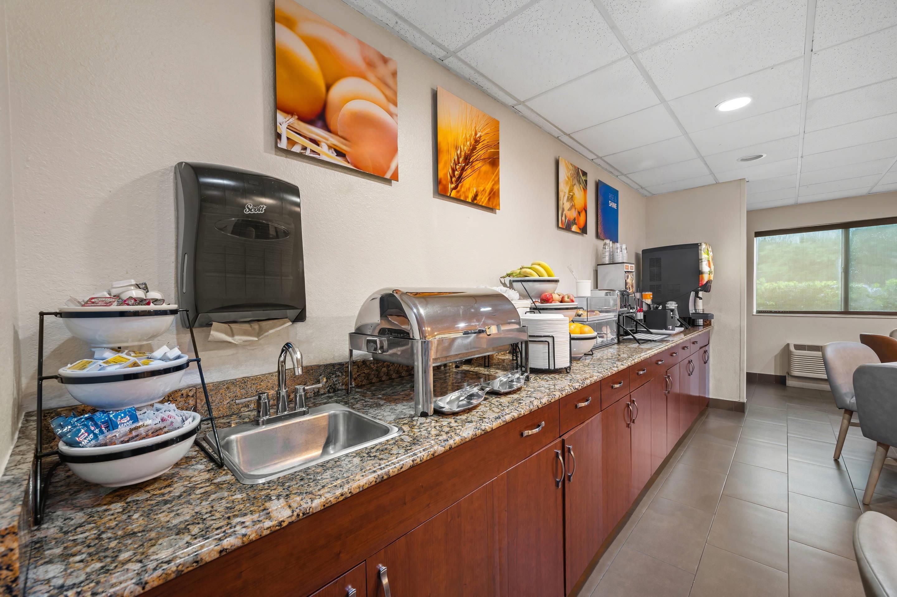 Espaço de refeições matinais com estação de autoatendimento, máquina de café e uma cesta de frutas.
