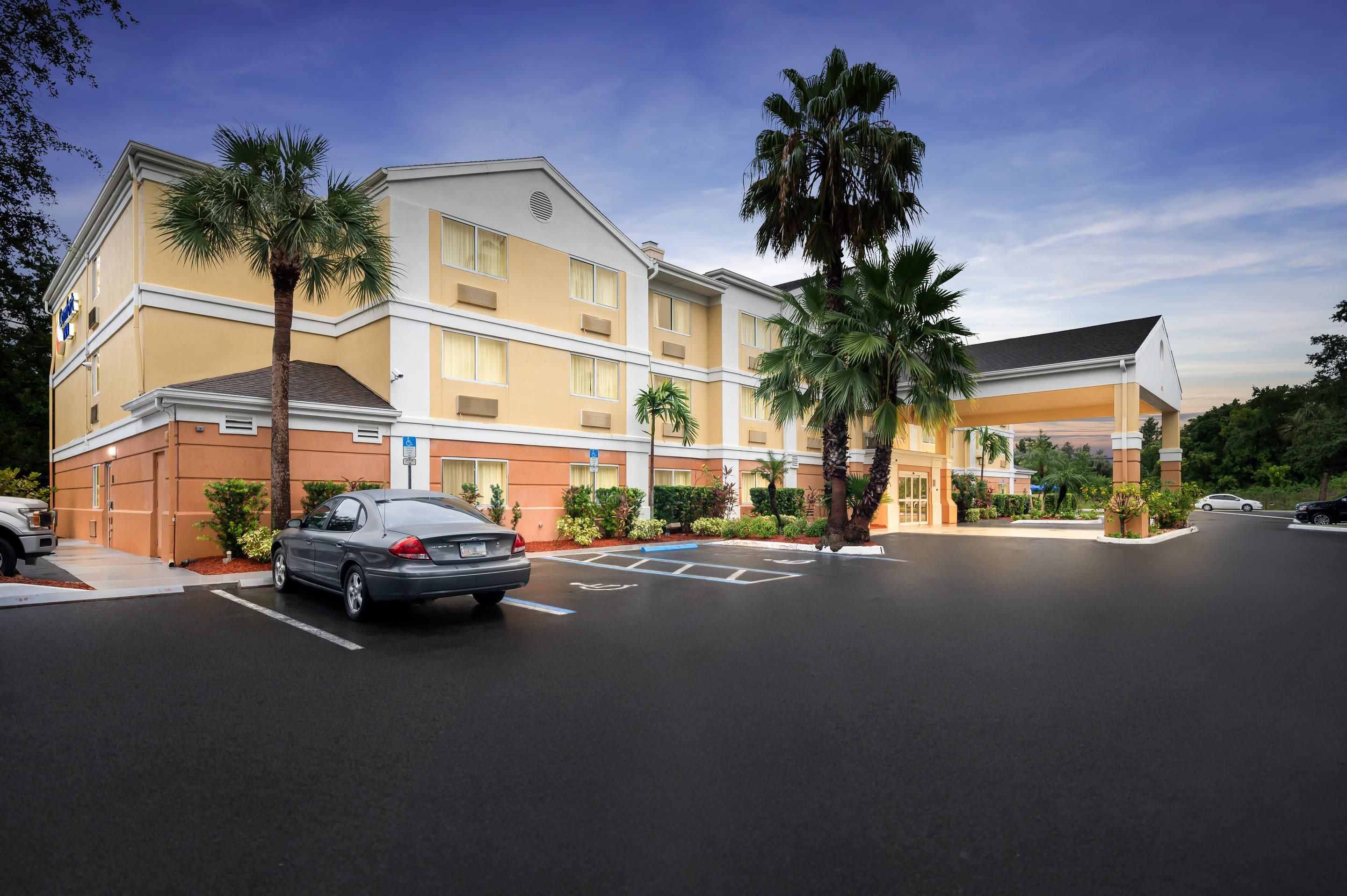A multi-story hotel with a parking lot, palm trees, and a clear sky.