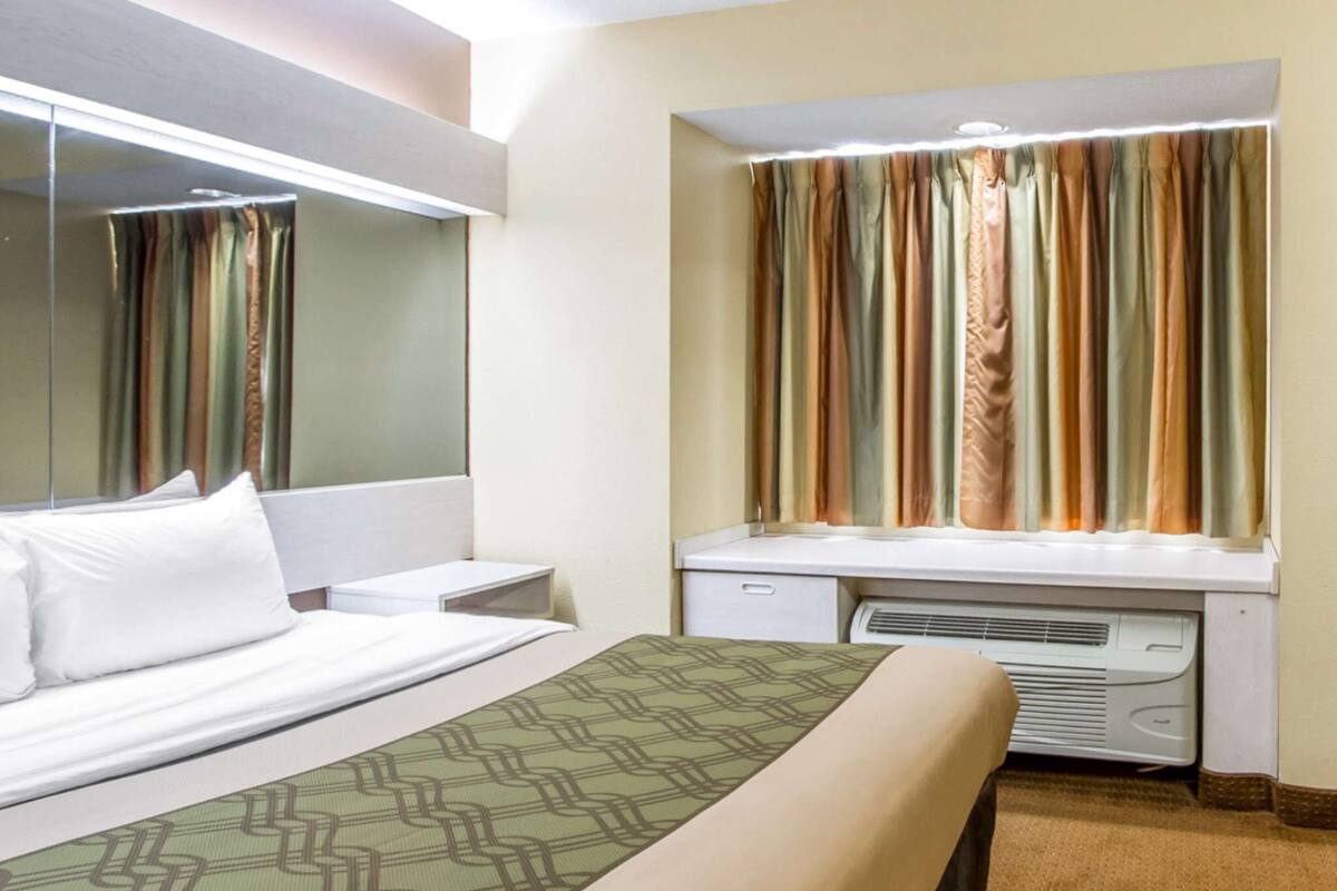 Una habitación de hotel con cama, escritorio, espejo y ventana con cortinas.