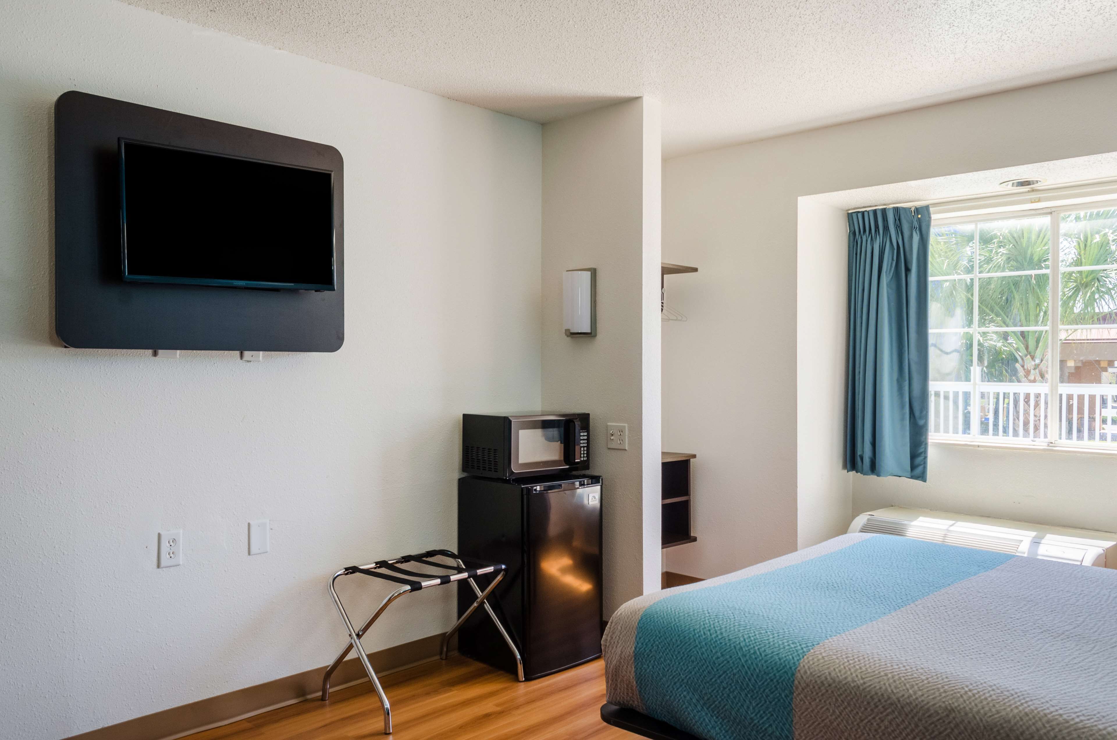 A hotel room with a bed, a TV mounted on the wall, a small refrigerator, a microwave, a window with curtains, and a wooden floor.