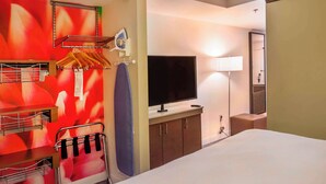 Habitación de hotel con cama, televisor, escritorio con lámpara, espejo y pared con un diseño abstracto en rojo.