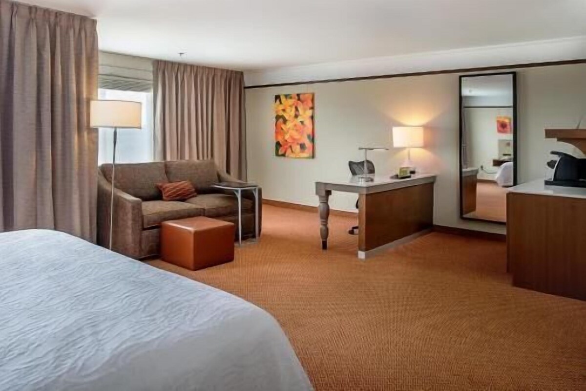 A hotel room with a bed, a sofa, a desk, a chair, a lamp, a painting, and a mirror.