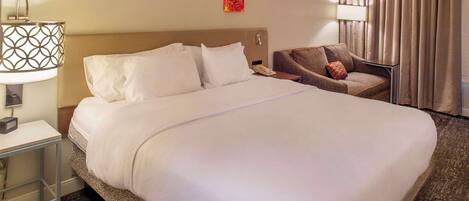 A hotel room with a large bed, a bedside table with a lamp, a chair with a cushion, a painting on the wall, and a window with curtains.
