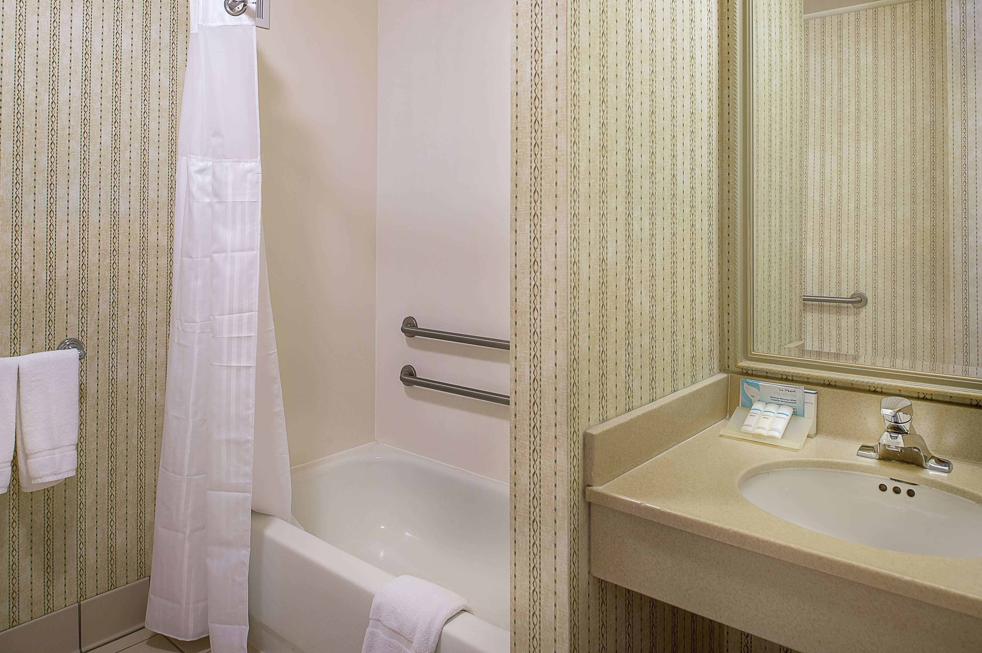 Ein Badezimmer mit Badewanne, Duschvorhang, Handläufer, Waschbecken mit Spiegel und Handtuchhalter.