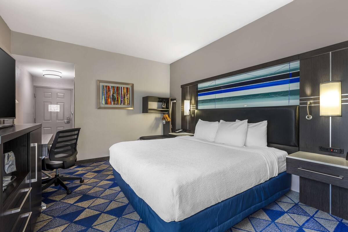 A hotel room with a large bed, a desk with a chair, a nightstand, a wall-mounted TV, and a patterned carpet.