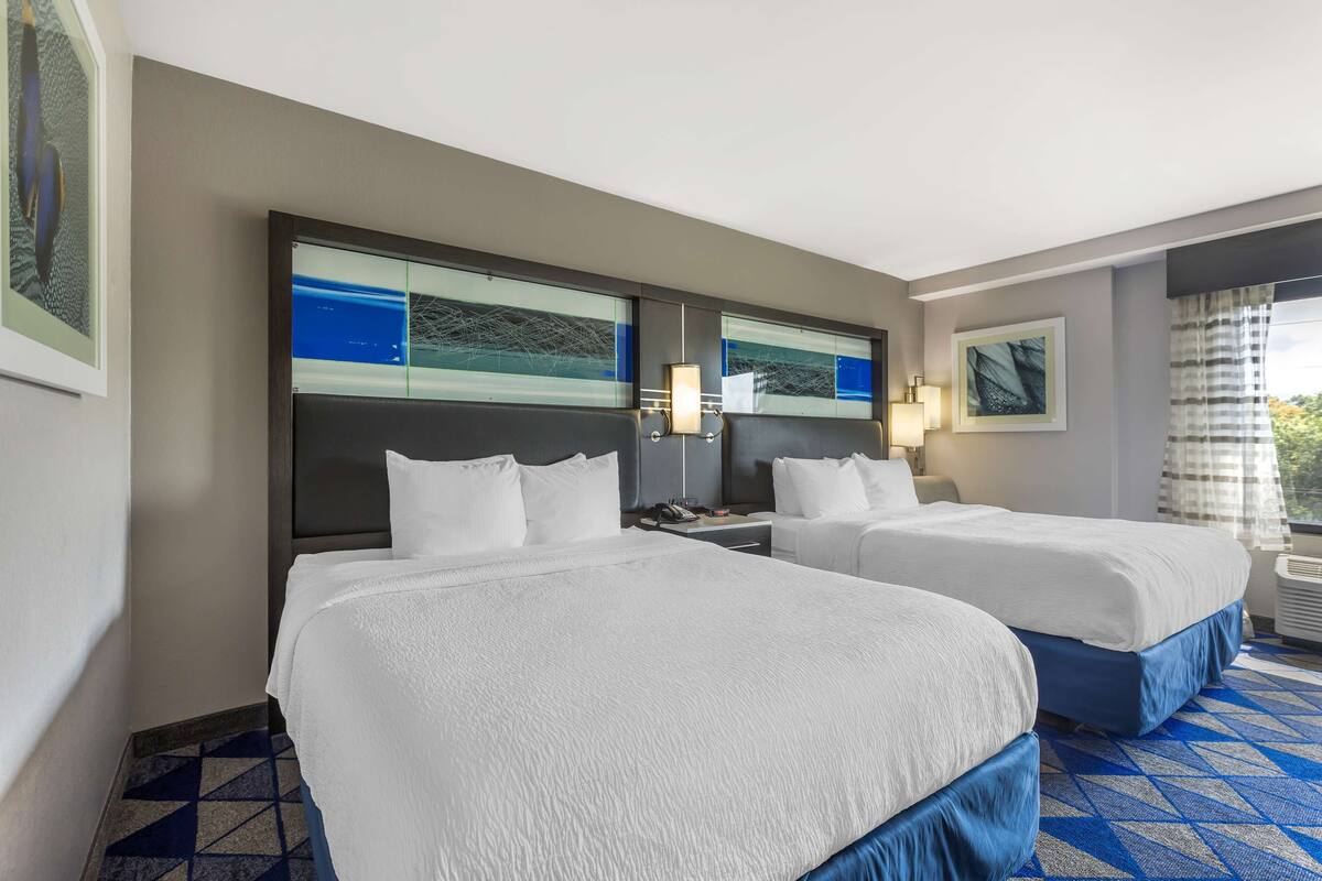 A hotel room with two beds, a large window with curtains, and artwork on the walls.