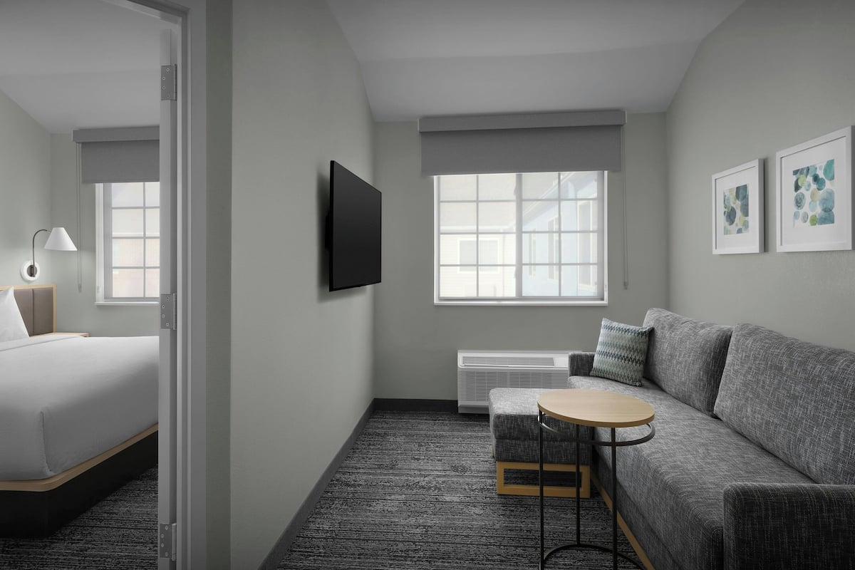 Una habitación de hotel moderna con una cama, un sofá gris, una mesita redonda pequeña, un televisor montado en la pared y una ventana con vistas.