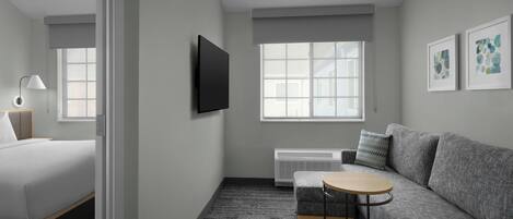 A modern hotel room with a bed, a gray sofa, a small round table, a TV mounted on the wall, and a window with a view.