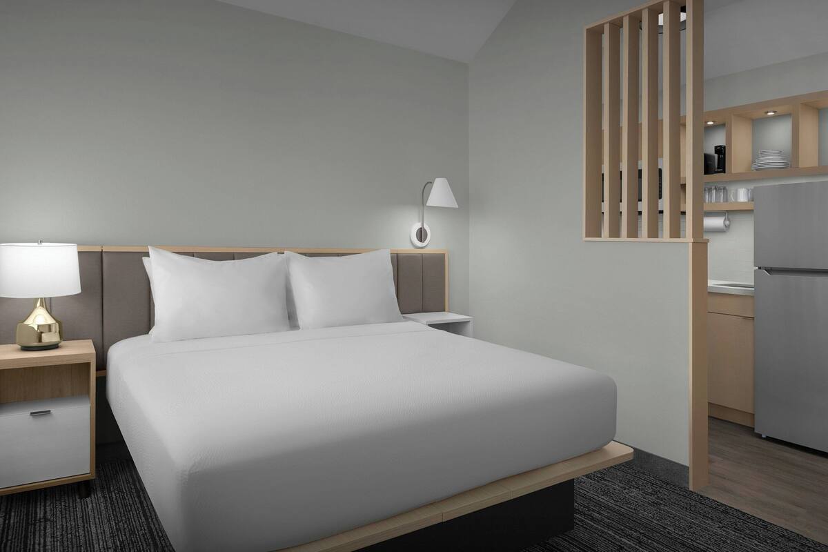 Habitación de hotel con cama, mesita de noche, lámpara y un rincón de cocina.