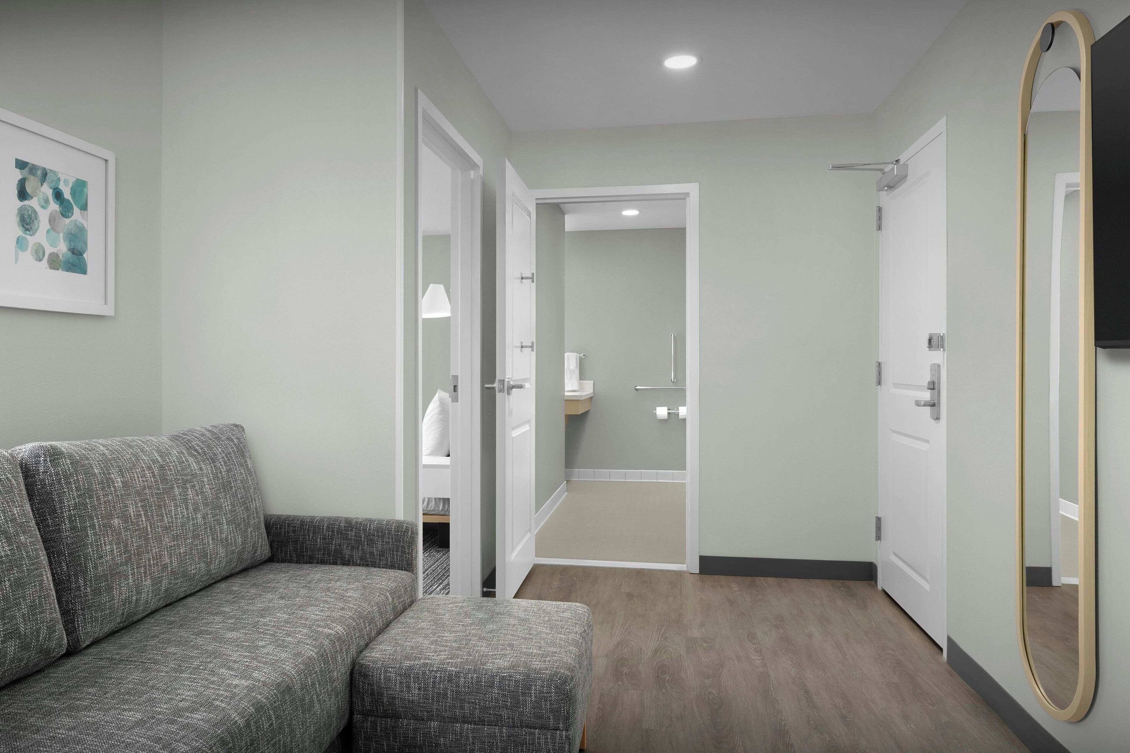 A modern hotel room with a grey sofa, a small ottoman, a wooden floor, and a bathroom visible through an open door.