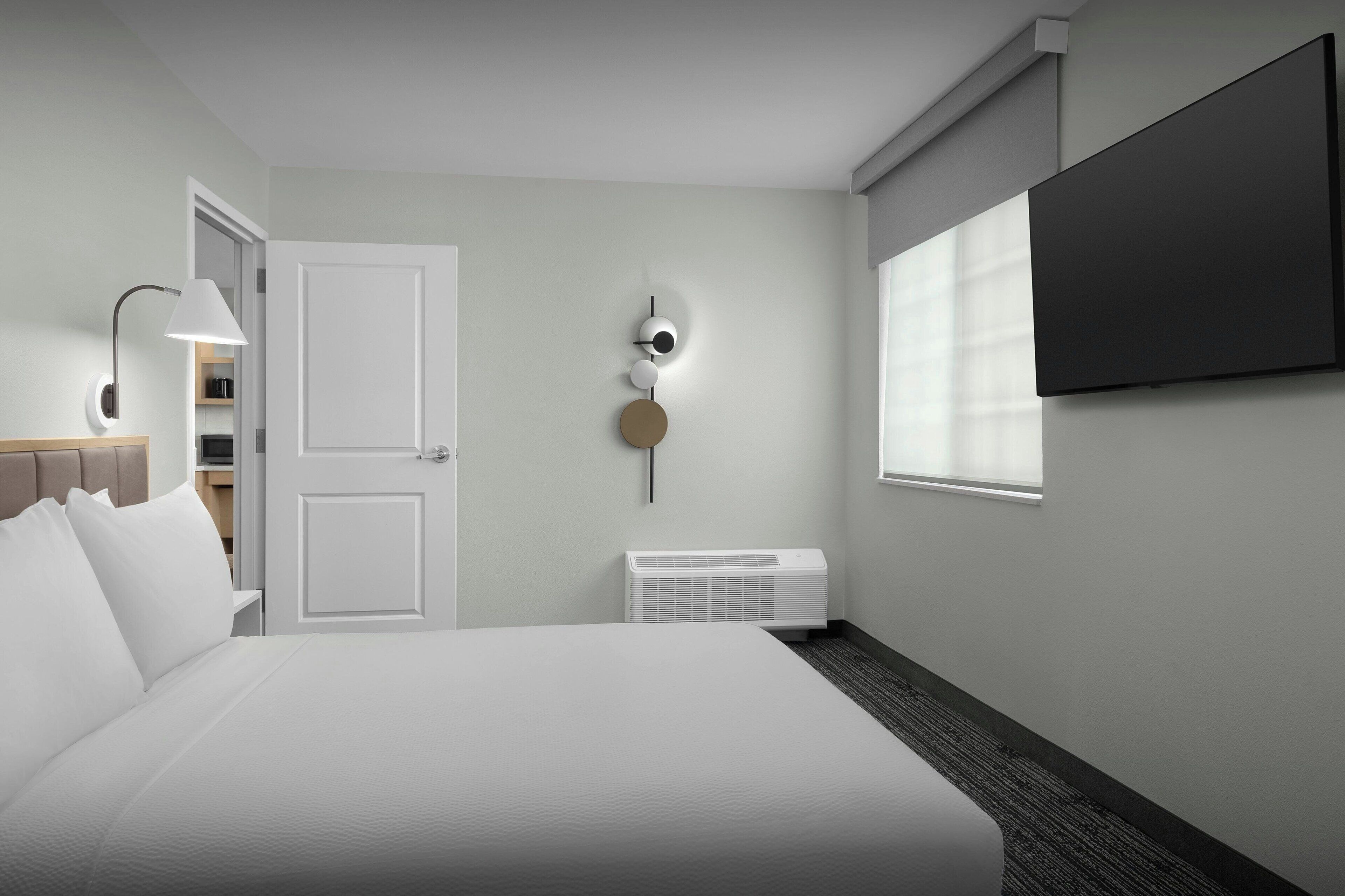 A hotel room with a bed, a TV, a wall-mounted light fixture, and a window.