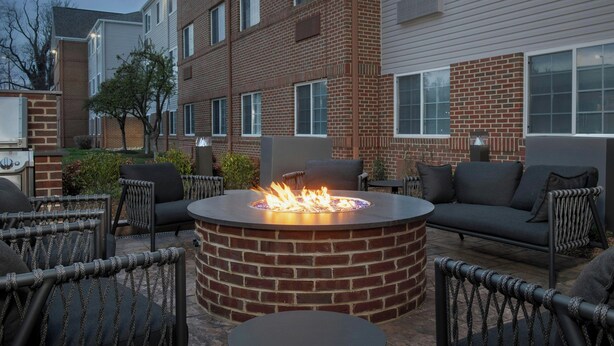 A circular fire pit surrounded by outdoor seating with a brick wall and a multi-story building in the background. - TownePlace Suites by Marriott Annapolis (Annapolis)