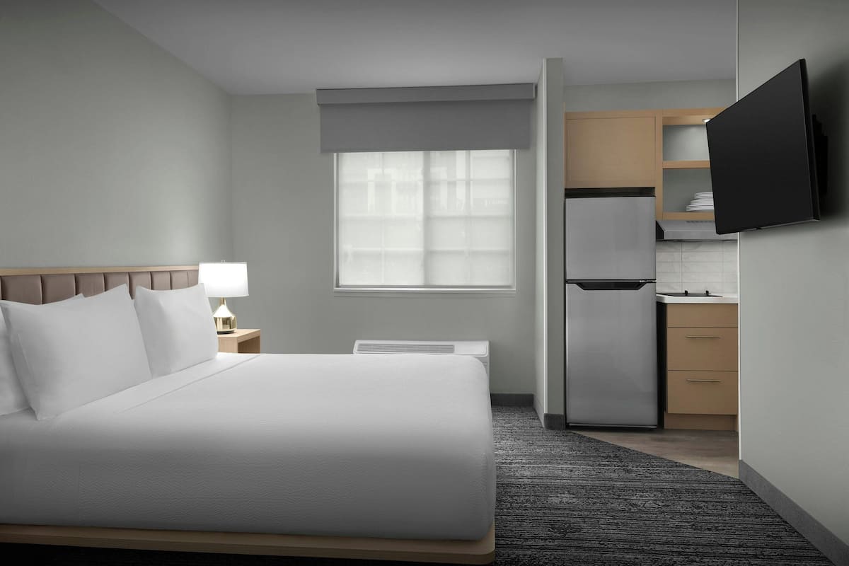 Una habitación de hotel moderna con una cama bien hecha, una mesita de noche pequeña con una lámpara, televisión integrada, una kitchenette compacta y una ventana con persianas.