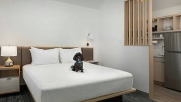 A hotel room with a bed, a nightstand with a lamp, a small dog on the bed, and a kitchenette area.