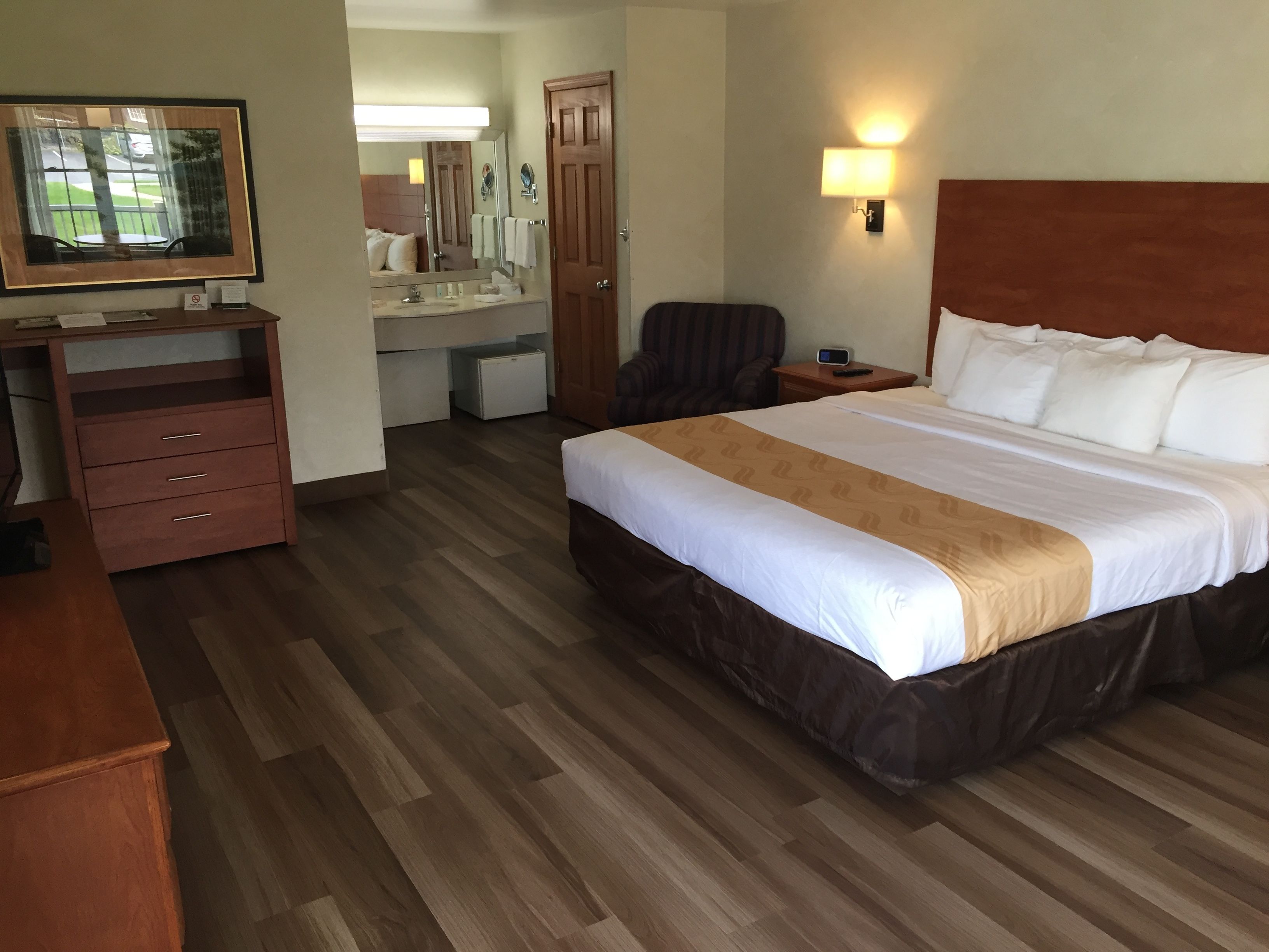 A hotel room with a bed, a desk, a chair, a dresser, a mirror, and a wall-mounted light.