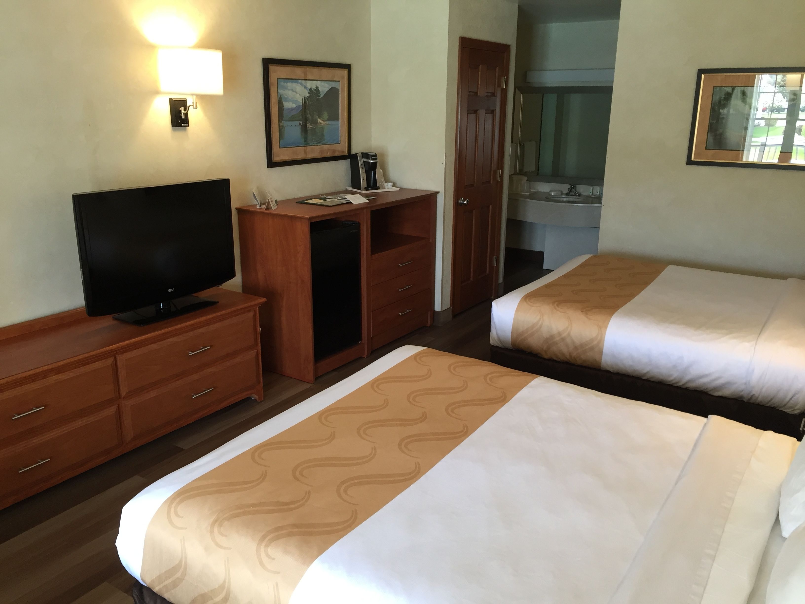 Habitación de hotel con dos camas, televisión, escritorio, armario y baño visible a través de una puerta abierta.
