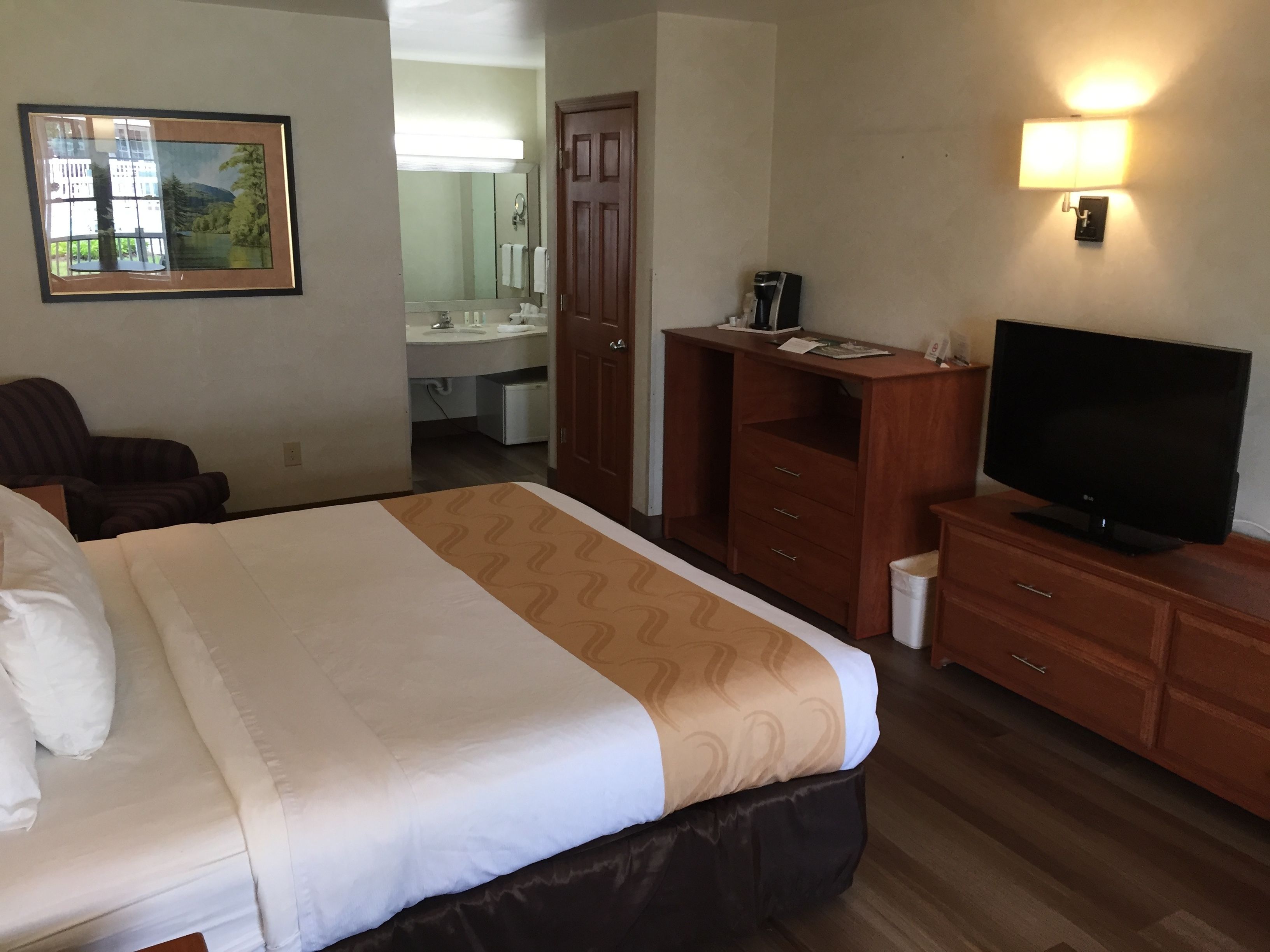 Quarto de hotel com uma cama grande, uma escrivaninha com um computador, televisão, espelho e banheiro visível por uma porta aberta.
