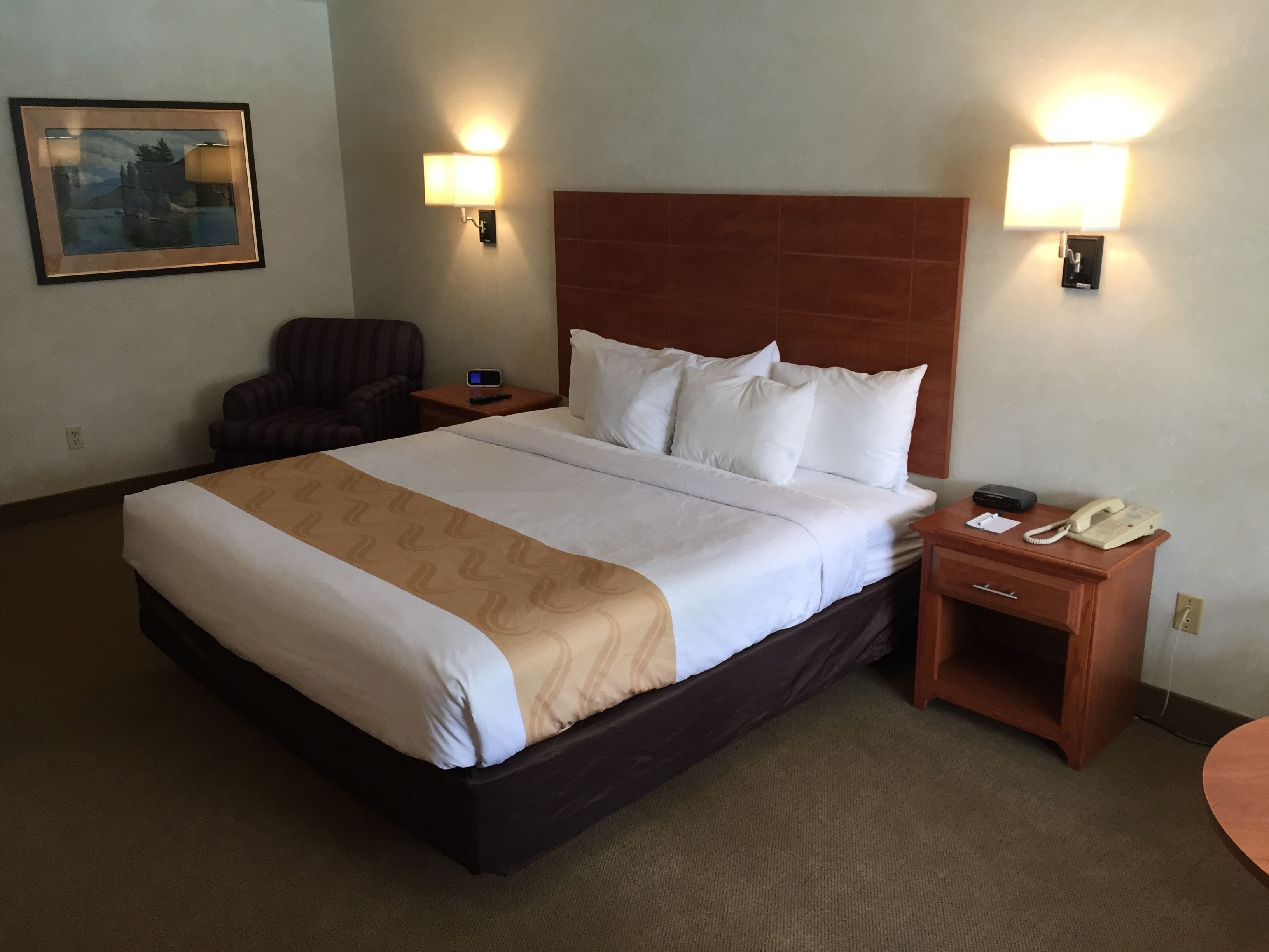 A hotel room with a large bed, two bedside lamps, a chair, a nightstand, a telephone, and a painting on the wall.