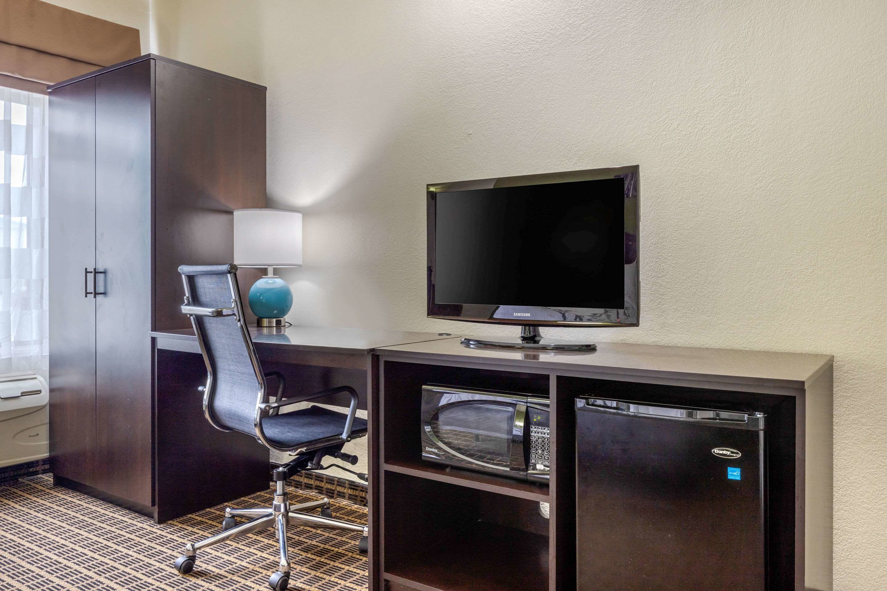 A hotel room with a dark wooden desk, a black office chair, a flat-screen TV, a microwave, and a mini-fridge.