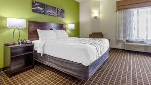 A hotel room with a bed, bedside tables, a nightstand, a lamp, a wall-mounted phone, a painting, a window with curtains, and an air conditioning unit. - Sleep Inn Orangeburg (Orangeburg)