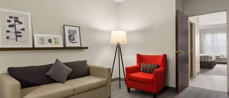 A hotel room with a beige sofa, a red armchair, a floor lamp, and framed artwork on the wall.