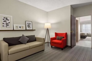 A hotel room with a beige sofa, a red armchair, a floor lamp, and framed artwork on the wall.