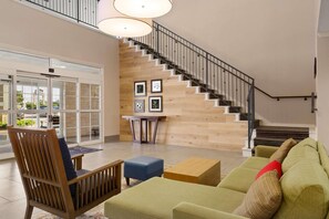 A spacious lobby with a staircase, wooden flooring, a seating area with a sofa and chairs, and a small table.