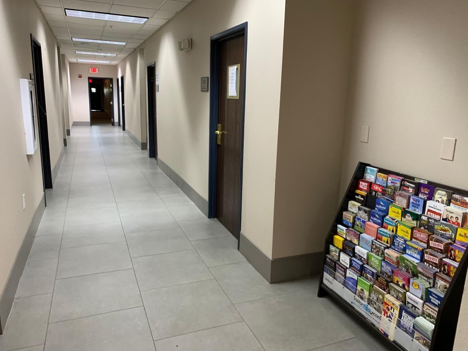 A hallway with a row of doors, a bulletin board with various pamphlets, and a tiled floor.