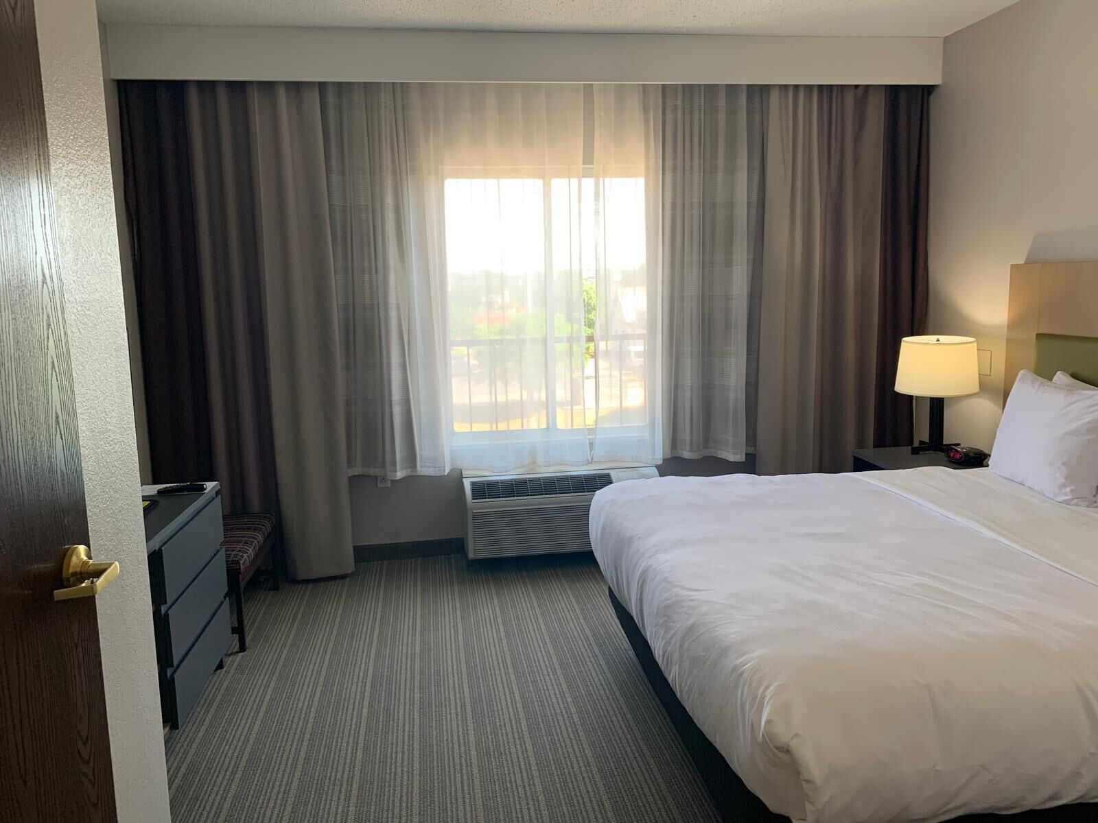 Habitación de hotel con cama, escritorio, silla, lámpara y ventana con cortinas.