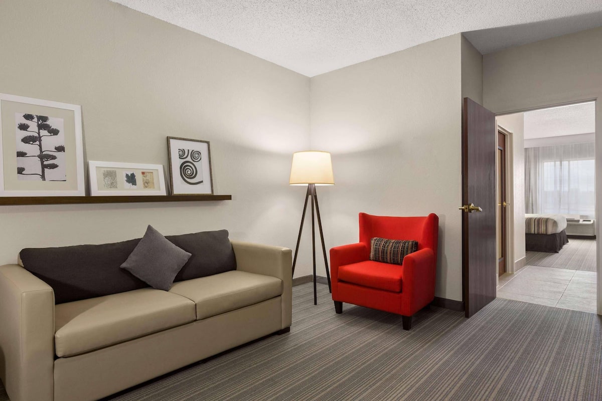 A hotel room with a beige sofa, a red armchair, a floor lamp, and framed artwork on the wall.