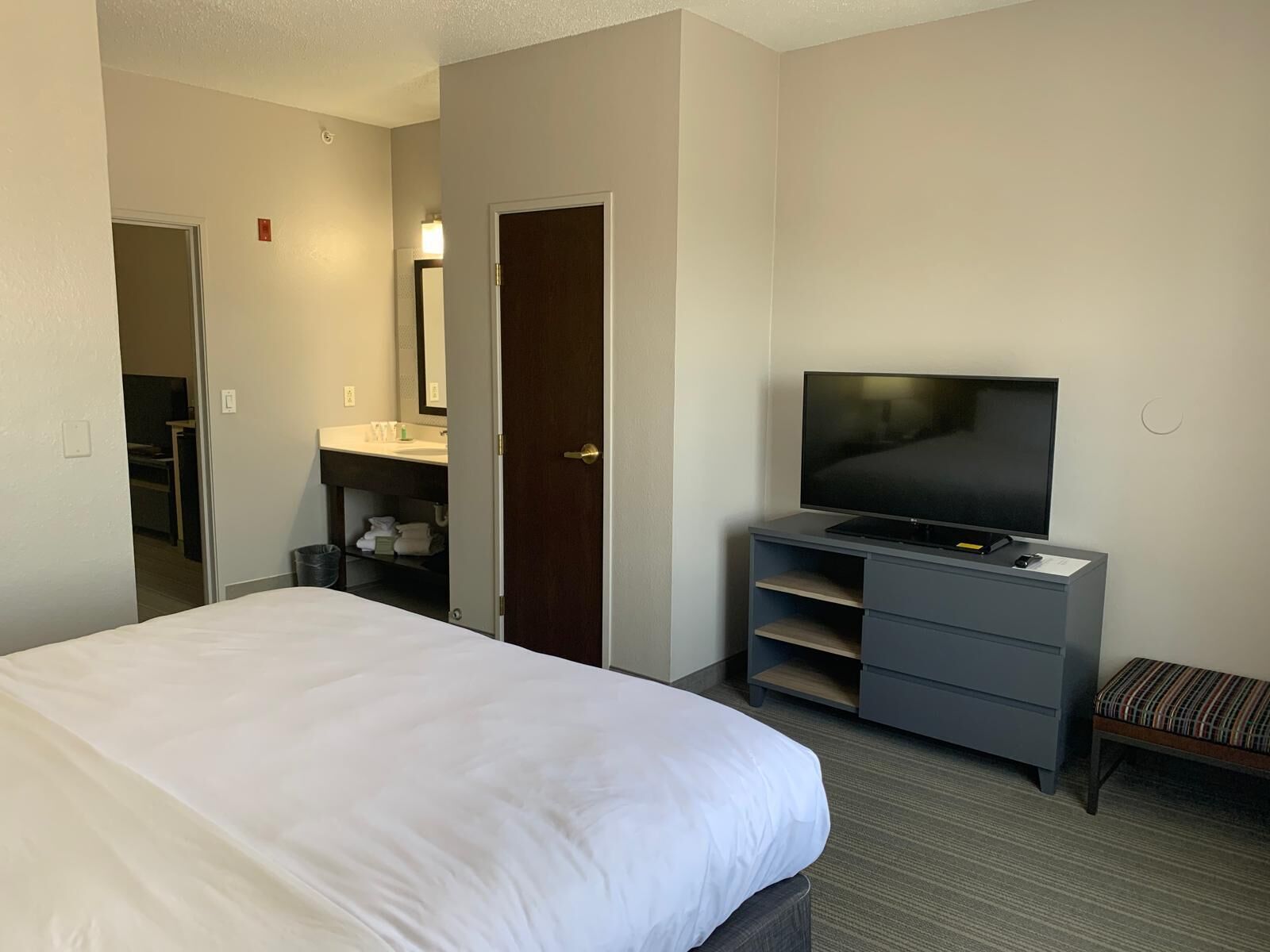 A hotel room with a bed, a television on a stand, a bathroom with a sink and mirror, and a door leading to another room.