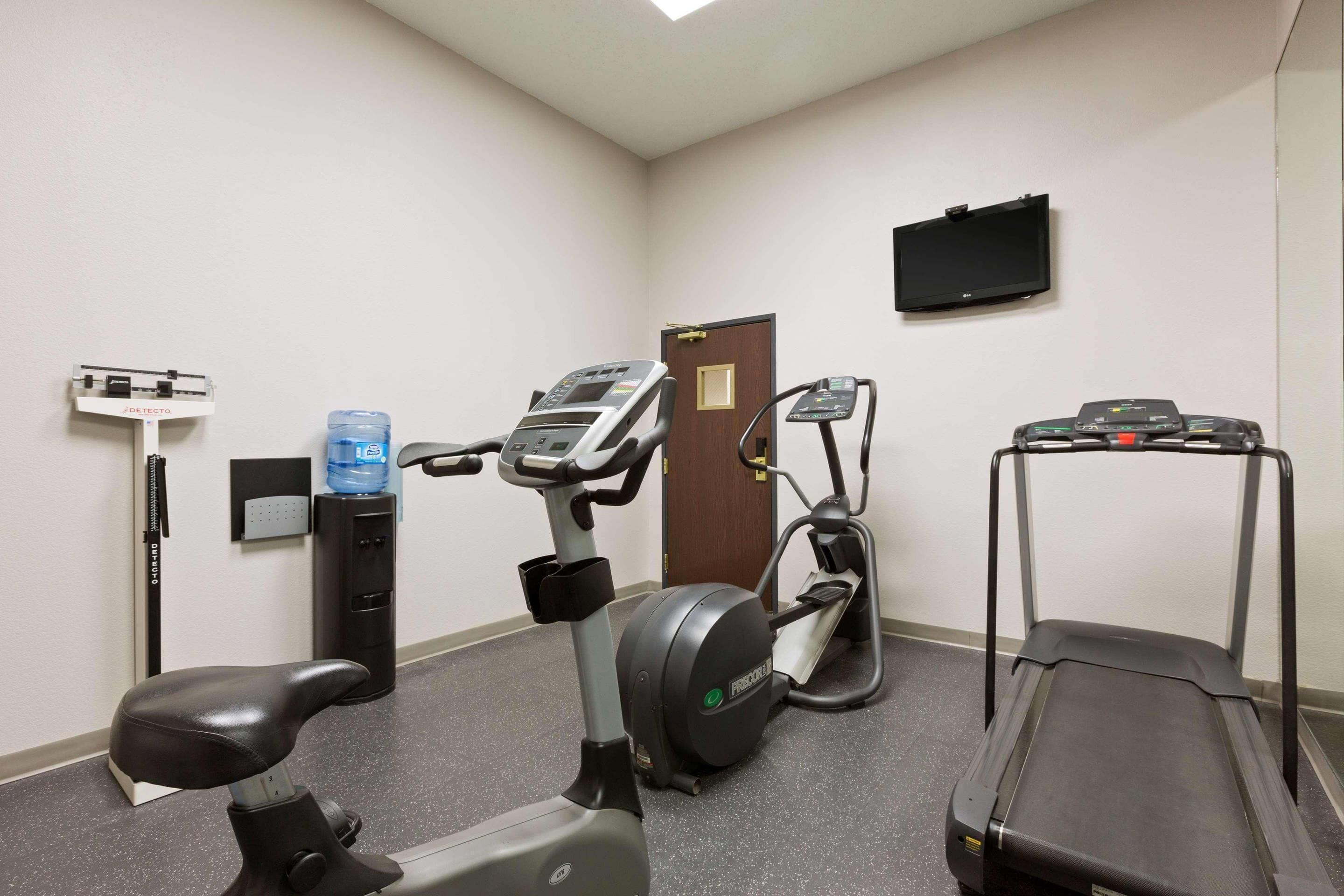A gym with stationary bikes, an elliptical, a treadmill, a water dispenser, and a TV mounted on the wall.