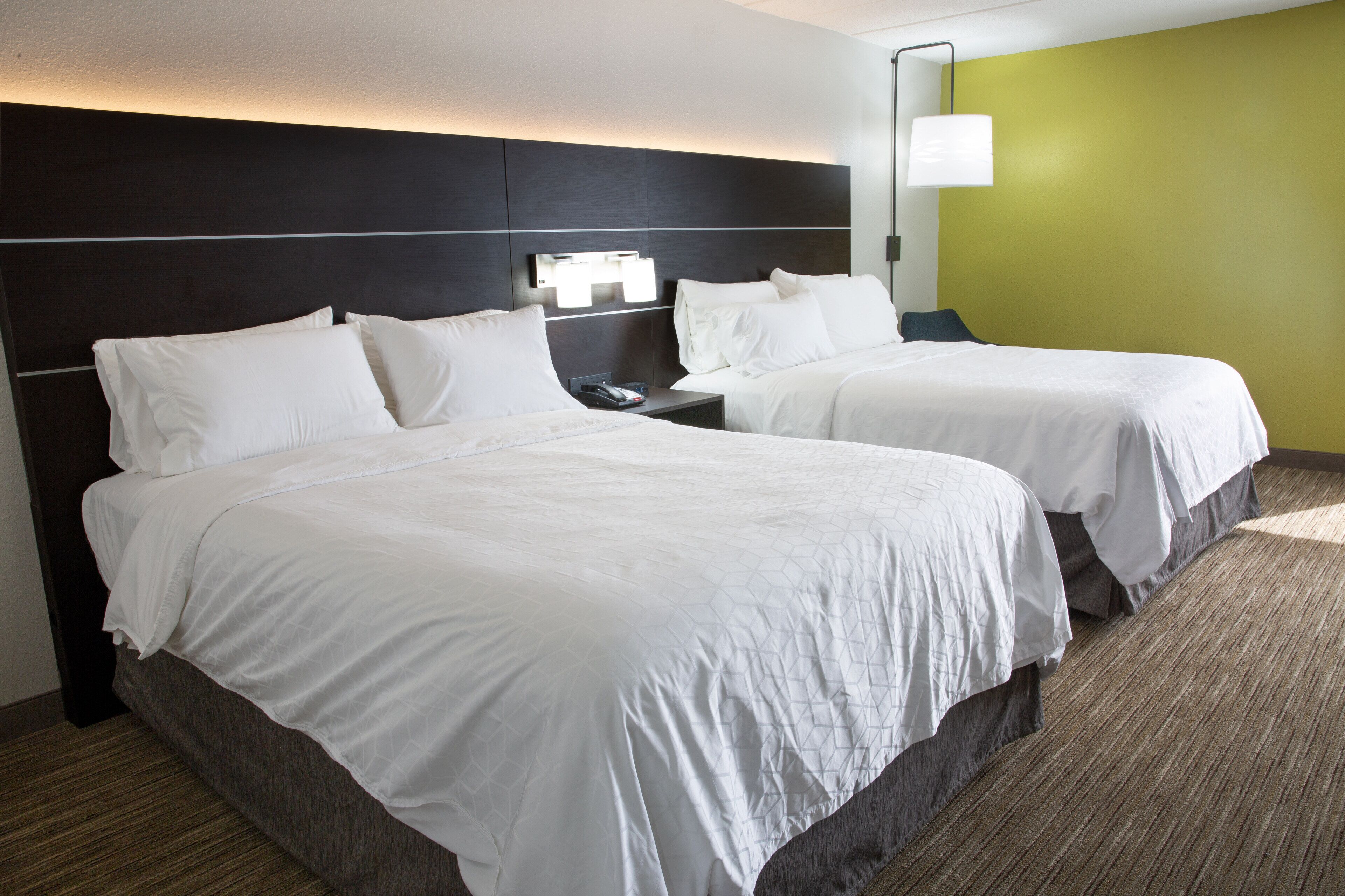 A hotel room with two beds, a nightstand, a lamp, and a wall-mounted light fixture.