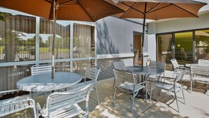 An outdoor patio area with round tables and metal chairs, shaded by large umbrellas, and a glass door leading to a covered walkway. - Hampton Inn Cincinnati-Northwest/Fairfield (Fairfield)